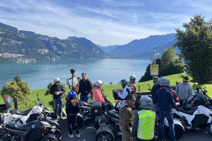 Group of motorcyclists with trikes by a lake with mountains in background.