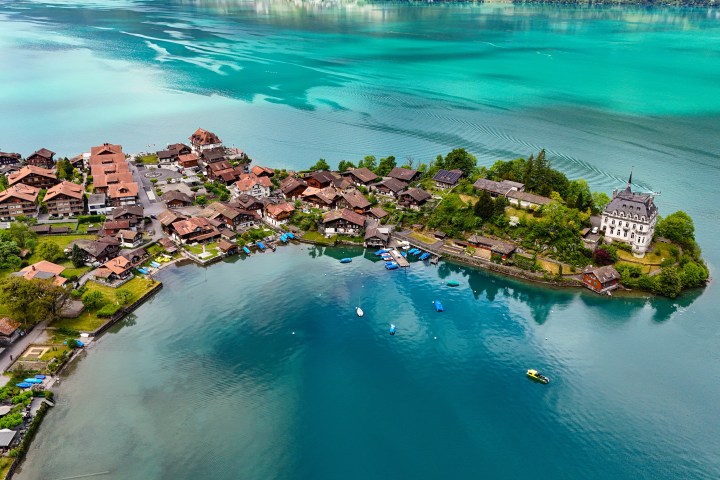 Aerial view of a lakeside village with houses and a mansion surrounded by turquoise water.