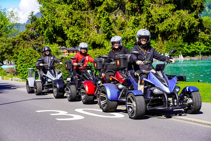Four people riding electric quadricycles on a lakeside road.
