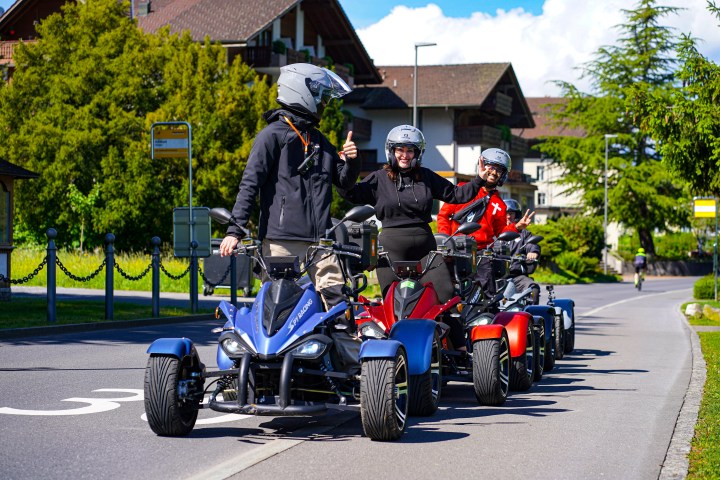 Four people on all-terrain vehicles parked on a road with buildings in the background.