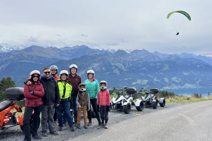 Group in helmets by three-wheelers with mountains and paraglider in background.