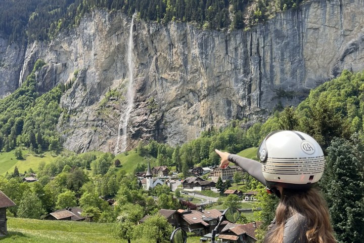 Person on a red scooter points at a waterfall with mountains in the background.