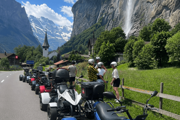 Line of quad bikes on road by mountains and waterfall, bright sky.